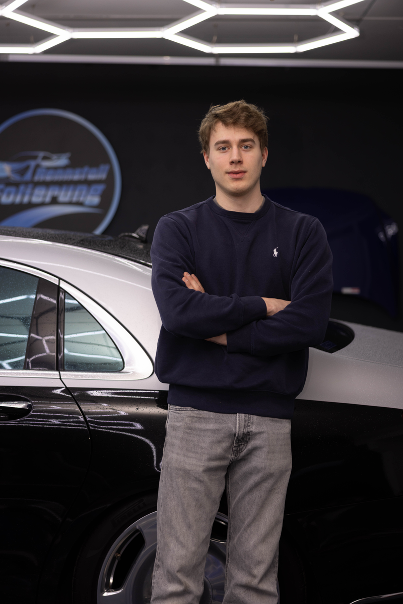 Junger Mann mit verschränkten Armen vor einem Auto unter Wabenleuchten